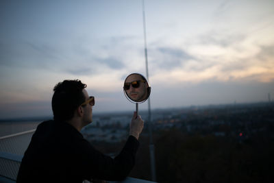 Side view of man holding mirror with his reflection against sky during sunset