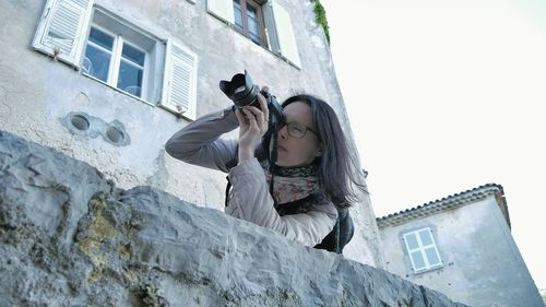 Low angle view of woman photographing while standing by buildings against sky