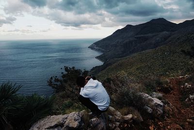 Rear view of man sitting on rock by sea against sky