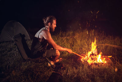 Side view of man sitting by campfire at night