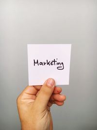 Cropped hand of person holding marketing text on adhesive note against gray background