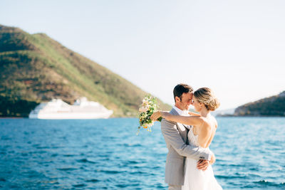 Side view of newlywed couple embracing while standing against sea