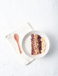 Top view of bowl with white yogurt topped with assorted cereals and sliced banana served for healthy breakfast on table with wooden spoon