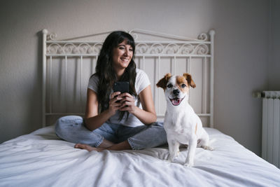 Smiling woman using smart phone sitting on bed at home