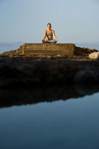 Peaceful female sitting on stone in padmasana and meditating with mudra gesture near sea during sundown