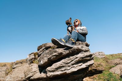 Low angle view of men and dog on rock against clear blue sky