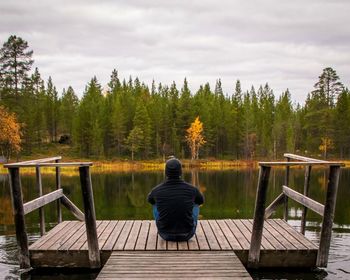 Rear view of man sitting on seat by lake against sky