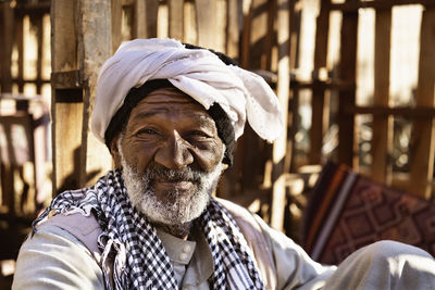 Aged nubian man in ethnic outfit sitting on carpet in yard and looking at camera in sunlight