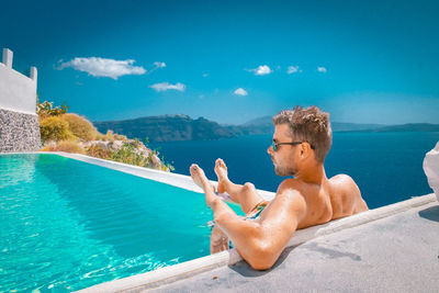 Shirtless man sitting by swimming pool against sea during sunny day