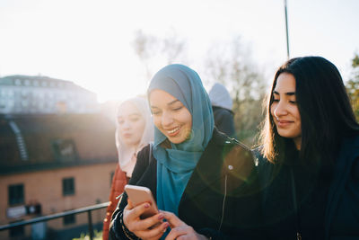 Back lit of smiling young woman using smart phone by female friends in city