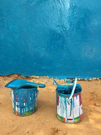 A wall painted in blue with two paint cans and two pencils dirty with blue paint on the sand floor 