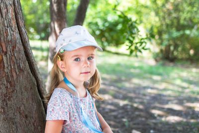 Little girl in a floral dress and hat leaning against a tree in the park