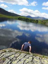 Rear view of man sitting on rock formation by lake against sky