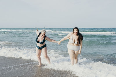 Positive curvy female friends in swimwear holding hands while walking in sea water during summer vacation