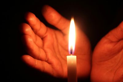 Cropped image of hands shielding burning candle against black background