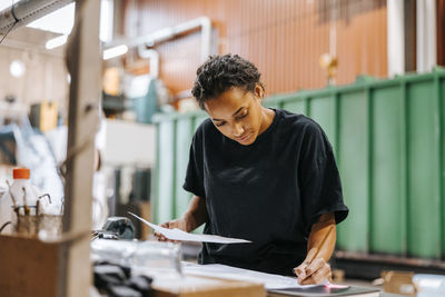 Female industry worker writing on blueprint while standing near table in manufacturing factory