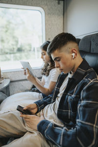 Boy wearing wireless in-ear headphones and using smart phone while sitting with sister in train