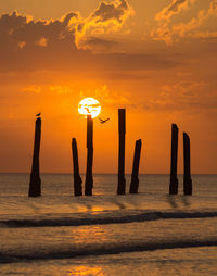 Silhouette wooden posts in sea against orange sky