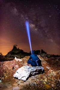 Rear view of man with illuminated flashlight sitting on rock at night