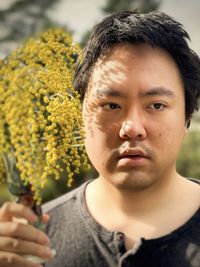 Close-up portrait of young man holding golden wattle flowers in vintage vase against trees.