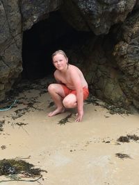 Portrait of shirtless boy crouching on sand against rock at beach