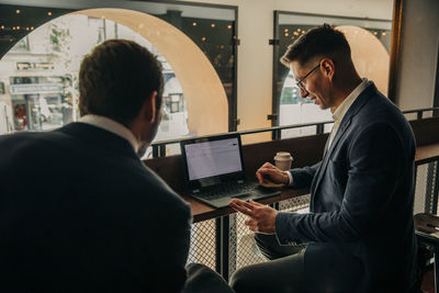 Businessman sharing laptop with male colleague while sitting in hotel lounge