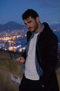 Portrait of handsome man using mobile phone against illuminated cityscape