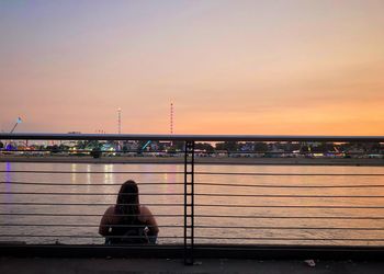 Man sitting on bridge against sky during sunset