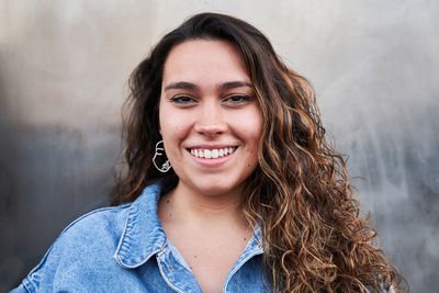 Optimistic young woman with long curly hair smiling and looking at camera while standing against gray metal wall on street of madrid, spain