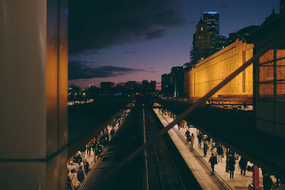 People on illuminated bridge by buildings against sky during sunset