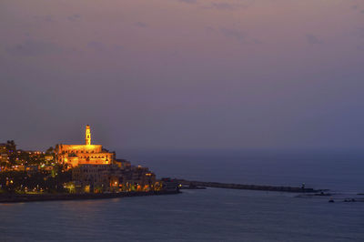 Illuminated buildings by sea against sky during sunset