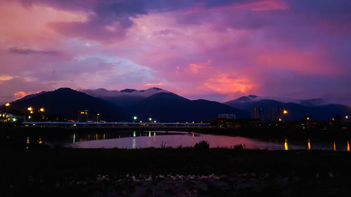 Illuminated city by lake against sky at sunset