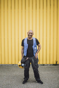 Full length of smiling bald construction worker standing against yellow metal wall