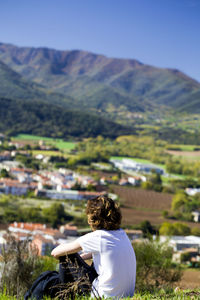 Young man sitting on field against town on sunny day