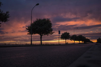 Silhouette trees by road against dramatic sky