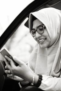 Smiling woman in wearing headscarf using phone while sitting in car