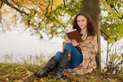 Full length portrait of woman reading book while leaning on tree trunk at lakeshore