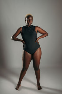 Serious african american curvy female in black bodysuit standing with her hands on her hips on gray background in studio