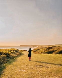 Rear view of woman walking on field against sky during sunset