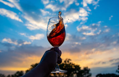 Hand holding glass against sky during sunset