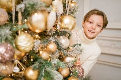 Happy guy in sweater at christmas tree decorates and looks out from behind it.