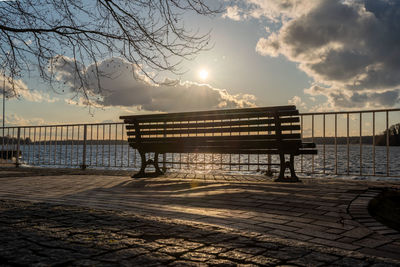 Silhouette bench on street by river against sky during sunset