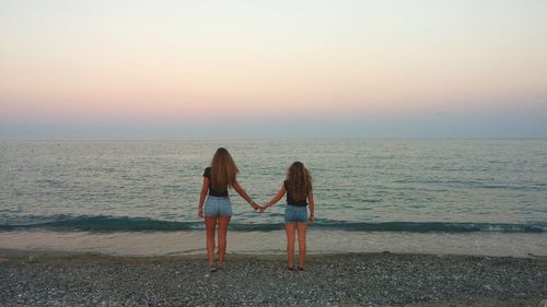 Rear view of friends holding hands while standing at sea shore against sky during sunset