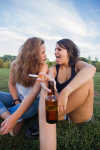 Portrait of smiling young woman holding beer bottle and cigarette while sitting with friend on field
