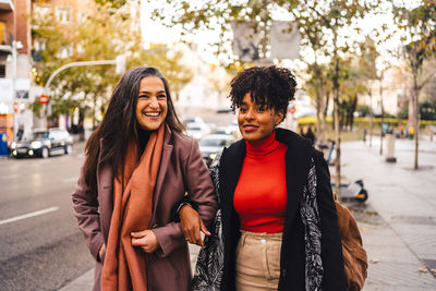 Cheerful lady holding arms black female friend in warm clothes while walking together on street pavement looking away