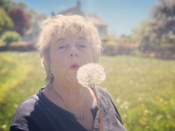 Portrait of senior woman blowing on dandelion seed standing on field