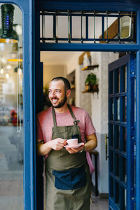 Positive bearded male barista in apron with cup of hot coffee in hands standing in doorway of modern coffee shop
