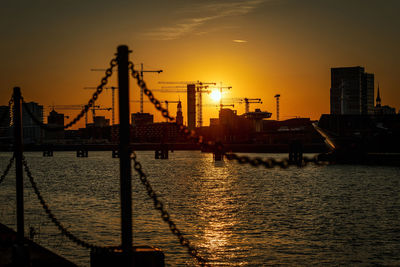 Silhouette cranes by river against sky during sunset