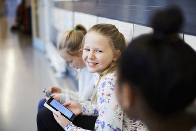 Smiling girl holding smart phone while looking at friend in school corridor