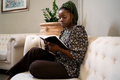 Side view of focused african american female with braids reading interesting book while resting on couch in modern living room at home